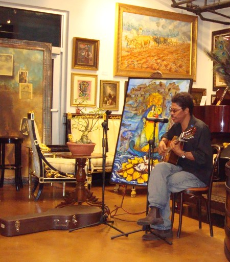 El trovador Alberto Cabrales interpretando sus canciones, junto al cuadro ÉXODO CACHITA, de Ignacio Pérez Vázquez, también presente en la Revista BRUJULAR DE MIAMI, y el Bonsai Babel, de Raúl Ortiz, padre de Melisa.