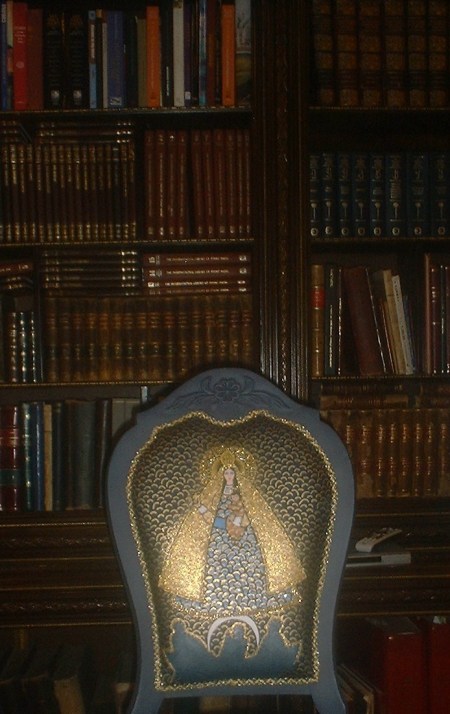 La maganífica estantería de la biblioteca del Centro de Arte CUBA 8, junto al mobiliario y la muestra de cuadros, cuyo centro es la Virgen de la Caridad del Cobre, nuestra Ochún. Mírela en el increible tapiz de esta silla.
